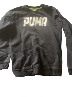Svart sweatshirt från Puma - Svart sweatshirt från Puma med stor vit logga på bröstet. Tröjan har rund halsringning, långa ärmar och ribbade muddar vid ärmslut och nederkant. Perfekt för en sportig och avslappnad stil.