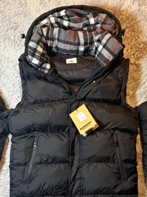 Svart dunjacka från Burberry - Snygg svart dunjacka från Burberry med avtagbara ärmar så den kan användas som väst. Storlek Xl sitter mer som m passar om du är 175-180. Helt ny oanvänd 