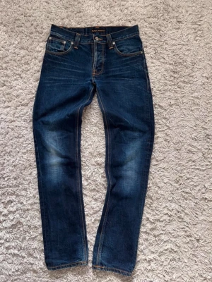 Mörkblå jeans från Nudie Jeans - Säljer ett par mörkblå jeans från Nudie Jeans med klassisk femficksmodell och kontrastsömmar. Jeansen har en slim fit passform. Tillverkade i denim av bomull. W30, L32.