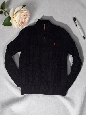 Svart kabelstickad tröja Polo Ralph Lauren - Svart kabelstickad tröja från Polo Ralph Lauren med halv dragkedja och hög krage. Tröjan har det klassiska röda broderade logotypen på bröstet och ribbstickade muddar. Perfekt för en stilren och avslappnad look.