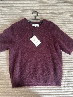 Vinröd stickad tröja från Gina Tricot  - Mysig vinröd stickad tröja från Gina Tricot med korta ärmar och rund hals. Tröjan är gjord i en fluffig alpackablandning som ger en mjuk känsla och trendig look. Perfekt att styla med jeans eller kjol för en avslappnad vibe.