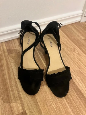 Svarta pumps med rem från Alley - Snygga svarta pumps från Alley med öppen tå, bred rem med volangdetalj framtill och justerbar ankelrem med silverfärgat spänne. Klackarna är medelhöga och skorna har en mjuk innersula. Materialet är mockaimitation.