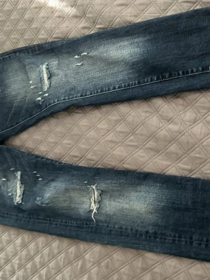 Mörkblå slitna jeans med patch - Säljer ett par mörkblå jeans med slitningar och en cool metallpatch med siffran 2 vid fickan. Jeansen har skinny fit och tydliga slitdetaljer på både lår och knän. Klassisk femficksmodell i denim, perfekt för en trendig streetstil.