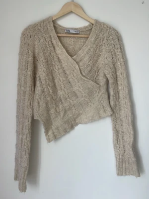 Beige stickad omlottopp från Zara - Trendig beige stickad omlottopp från Zara med kabelstickat mönster och asymmetrisk nederkant. Långärmad modell med v-ringning och ribbade muddar ❣️