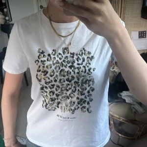 Vit t-shirt med leopardmönster från Zara - Säljer en vit t-shirt från Zara med ett stort leopardmönster i svart, beige och guld på framsidan. T-shirten har rund halsringning och korta ärmar. Skriv vid frågor och funderingar!