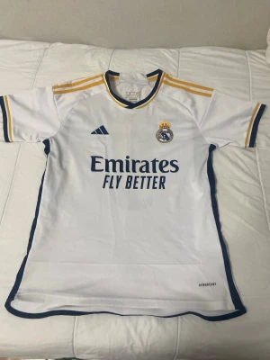Real Madrid matchtröja Adidas vit - Vit Real Madrid matchtröja från Adidas med gula och mörkblå detaljer. Tröjan har klubbmärke på bröstet, Adidas-logga och sponsortryck 'Emirates Fly Better' framtill. Tillverkad i lätt funktionsmaterial Aeroready, perfekt för fotbollsträning. Jätte fin bellingham tröja. Kolla in min profil du kanske gillar något. Fixar bra priser 