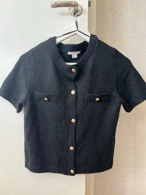 Svart kortärmad stickad kofta Gina Tricot - Kortärmad svart stickad kofta från Gina Tricot med rund halsringning och dekorativa guldknappar framtill. Två små fickor med knappdetaljer på bröstet. Ribbstickade kanter och mjukt stickat material ger en trendig look. Storleken är i Young från Gina Tricot, motsvarar XS i dam 