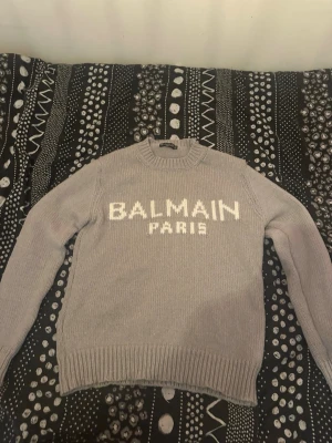 Grå stickad tröja från Balmain - Snygg grå stickad tröja från Balmain Paris med vit logotyp framtill. Tröjan har rund halsringning och ribbstickade muddar vid ärmslut och nederkant. Perfekt för dig som gillar exklusiva märken och stilren design. Säljer denna pga att den är lite för stor på mig antar att den är stor i storlek. Skickar fler bilder ifall de behövs!