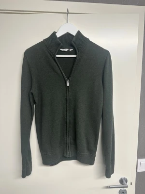 Mörkgrön ziptröja med hög krage - Snygg mörkgrön ziptröja med hög ribbad krage och hel dragkedja framtill. Tröjan har långa ärmar och ribbade muddar vid ärmslut och nederkant. Perfekt för lager på lager och enkel att matcha med olika outfits.