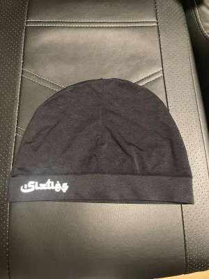 Svart sixties mössa - Säljer en svart, tunn och mjuk mössa med vitt tryck på sidan. Mössan har enkel design utan mönster och är tillverkad i stretchigt tyg som sitter skönt på huvudet. Perfekt för dig som vill ha en stilren och enkel accessoar till vardags.