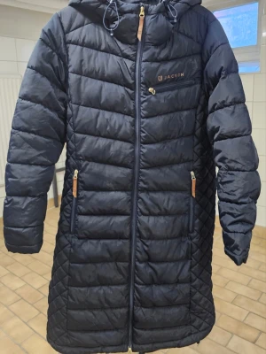 Svart lång dunjacka från Jacson - Snygg svart lång dunjacka från Jacson med huva och dragkedja framtill. Jackan har två framfickor med dragkedja och en bröstficka, samt quiltade detaljer på sidor och ärmar. Perfekt för kalla dagar och riktigt skön att bära.