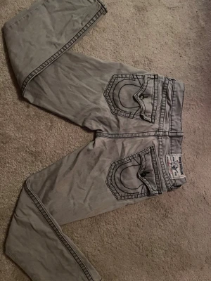 Grå True Religion jeans med kontrastsömmar - Säljer ett par grå jeans från True Religion med tydliga svarta kontrastsömmar och stora bakfickor med lock och knapp. Klassisk logga på bakfickan och patch i midjan. Snygg streetstil och unik design som sticker ut.