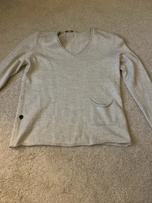 Ljusgrå tröja från House of Lola - Mjuk och stilren ljusgrå tröja från House of Lola med v-ringning och lång ärm. Tröjan har en liten ficka framtill och en diskret knappdetalj vid nederkanten. Perfekt för en avslappnad och enkel look.