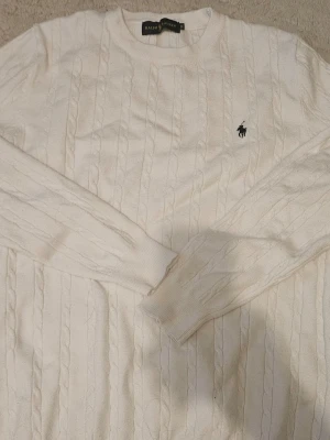 Vit kabelstickad tröja från Ralph Lauren - Kabelstickad vit tröja från Ralph Lauren med klassisk rund halsringning och broderad marinblå logga på bröstet. Tröjan har långa ärmar och ribbade muddar. Perfekt för en stilren och tidlös look.