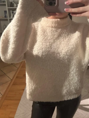 Krämvit fluffig stickad tröja - Mysig krämvit stickad tröja med fluffig textur och hög hals. Tröjan har långa ärmar och en avslappnad passform, perfekt för kalla dagar. Materialet känns mjukt och gosigt mot huden. Enkel och stilren design utan synliga loggor eller mönster.