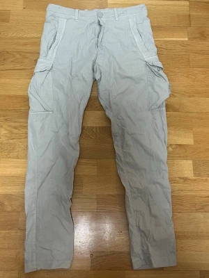 Ghost Edition: Sandfärgade cargobyxor från Stone Island - Ghost Edition: Säljer ett par sandfärgade cargobyxor från Stone Island med klassiska stora fickor på sidorna och broderad logga på ena benet. Byxorna har raka ben, bälteshällor och är tillverkade i ett lätt bomullsmaterial. Perfekta för en avslappnad och trendig stil.