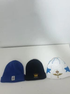 Tre coola mössor i olika färger - Säljer tre mössor: en blå ribbstickad mössa från Junkyard med uppvikt kant, en svart mössa med guldkrona och texten 'Prince', samt en vit mössa med blå stjärnor och broderat emblem framtill. Perfekta accessoarer för att hålla stilen när det är kallt.