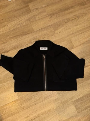 Svart zip cardigan från Samsøe Samsøe - Stilren svart cardiganjacka från Samsøe Samsøe med dragkedja framtill och klassisk krage. Jackan är i ett mjukt stickat material och har långa ärmar samt en bröstficka för extra detalj. Perfekt för en clean och modern look.