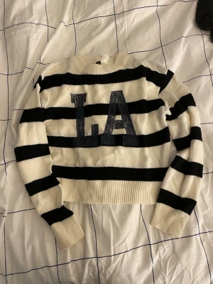 Randig stickad tröja med LA-tryck - Säljer en vit och svart stickad tröja med breda horisontella ränder och stort blått 'LA'-tryck på bröstet. Tröjan har rund halsringning, långa ärmar och ribbade muddar. Perfekt för dig som gillar streetstyle och vill ha en cool statement-piece.