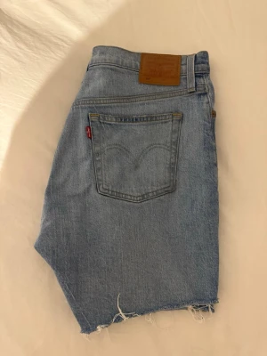 Levi's 501 ljusblå jeansshorts - Ljusblå jeansshorts från Levi's modell 501 med klassisk femficksdesign och råa, fransiga benslut. Ikonisk läderpatch bak i midjan och röd Levi's-tagg på bakfickan. Perfekta för en avslappnad stil.