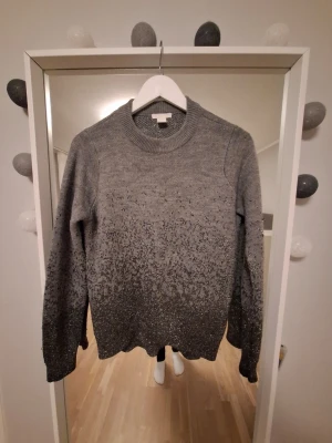 Grå stickad tröja med glittereffekt - Snygg grå stickad tröja med glitterdetaljer som ger en skimrande effekt, perfekt för dig som vill sticka ut lite extra. Tröjan har rund halsringning, långa ärmar och ribbade muddar vid ärmslut och nederkant. Materialet känns mjukt och bekvämt.
