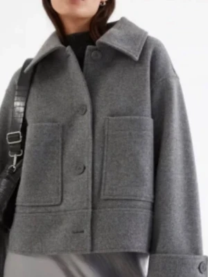 Grå ulljacka från KAPPHAL  - Grå jacka i bomull från Kappahl, nästan helt oanvänd, stora framfickor och bred krage. Jackan har knäppning framtill med stora knappar och uppvikta ärmslut. 