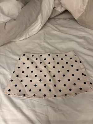 Prickig kortkjol i beige och svart - Supersöt kortkjol i beige med svarta prickar. Kjolen har en rak passform och är perfekt för dig som gillar lekfulla mönster. Tillverkad i ett mjukt och lätt material som känns skönt mot huden. Ingen synlig dragkedja eller knappar.