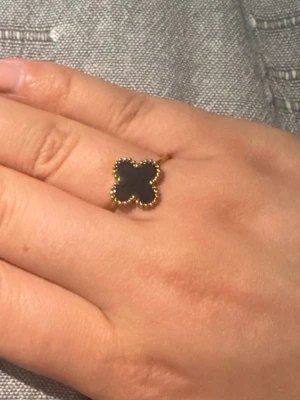 Fyrklöver ring i svart - Snygg guldfärgad ring med en svart klöverformad detalj. Ringen har små pärlliknande kanter runt klövern och en tunn, elegant ringband. Perfekt för dig som gillar unika och trendiga accessoarer.