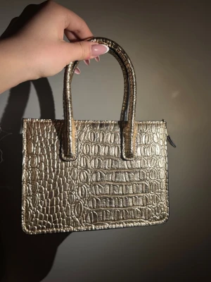 Guldig handväska med krokopräglat mönster - Snygg guldig handväska i fyrkantig modell med krokopräglat mönster. Väskan har två stadiga handtag och dragkedja upptill. Materialet är konstläder med metallic-finish som ger en lyxig känsla. Perfekt accessoar för att lyfta din outfit.