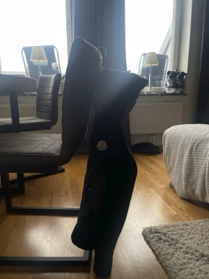 Svart cardigan från Moncler - Säljer en svart cardigan från Moncler med quiltad kropp och stickade ärmar. Jackan har huva, två framfickor med knappar och en extra ficka på ärmen. Klassisk Moncler-logga på bröstet och dragkedja framtill. Perfekt för kalla dagar.