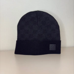 Svart ribbstickad mössa med patch - Snygg svart mössa med ribbstickad uppvikt kant och diskret patch framtill. Klassisk form som sitter skönt på huvudet och passar till de flesta stilar. Materialet ser ut att vara mjukt och värmande, perfekt för kyliga dagar.