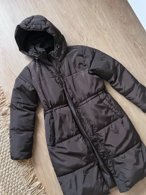 🤎 Y2K Chokladbrun Long Puffer Coat – Maxi Dunjacka med luva, Cubus - strl XS - Statement alert 🚨 Den perfekta chokladbruna longline puffer-jackan med clean silhuett och äkta Y2K energi. Maxilängd, oversized känsla och fluffig som ger den där effortless 00’s looken.  Perfekt att styla med low rise jeans, uggs eller chunky sneakers för full 2000s vibe <3  ✔️ Trendig chokladbrun nyans ✔️ Lång modell (maxi) ✔️ Dragkedja framtill ✔️ Huva ✔️ Mysig & varm – perfekt för höst/vinter/vår