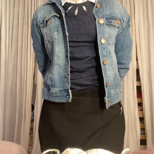 Klassisk blå jeansjacka med knappar - Säljer en klassisk blå jeansjacka med bronsfärgade knappar och två bröstfickor. Jackan har en tidlös passform och är tillverkad i slitstarkt jeansmaterial. Perfekt att slänga över en tröja för en cool och avslappnad look. Har en liten fläck, men såna knappt. På namnlappen har det redan skrivits ett namn men det kan täckas över med en klisterlapp!