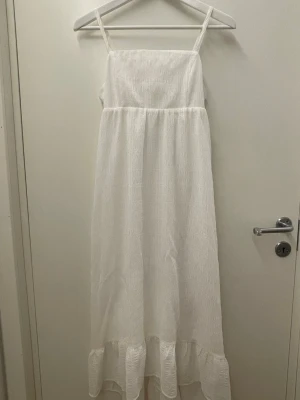 Vit långklänning från H&M Divided - Luftig vit långklänning från H&M Divided med smala axelband och volang nertill. Klänningen har en rak passform och är tillverkad i ett lätt, strukturerat tyg av polyester och bomull. Perfekt för varma dagar och somriga tillfällen.