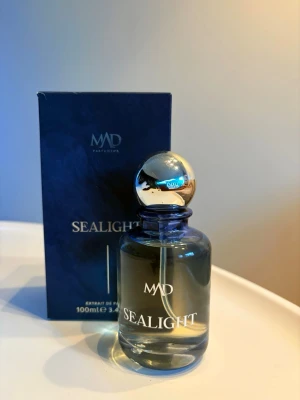  MAD Parfumeur - Sealight 100/100ml - Sealight från MAD Parfumeurs är en elegant parfym i en rund glasflaska med silverfärgad klotformad kork. Flaskan har en stilren design med vit text och rymmer 100 ml. Förpackningen är mörkblå och matchar flaskans exklusiva känsla.