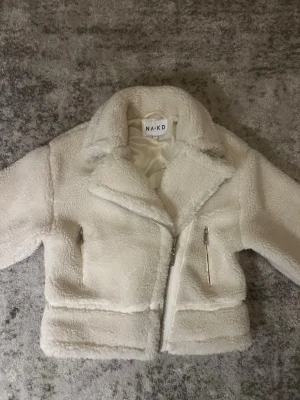 Vit teddyjacka från NA-KD - Supermjuk och fluffig teddyjacka i offwhite från NA-KD i storlek 36. Perfekt inför våren! Jackan har bred krage, två fickor med dragkedja och en snedställd dragkedja framtill. Skriv vid frågor eller fler bilder!🩷