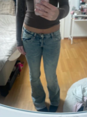 Blå bootcut jeans med strassdetaljer - Säljer ett par ljusblå bootcut jeans med låg midja och snygga strassdetaljer på bakfickorna. Jeansen har dekorativa knappar och klassisk femficksmodell. Perfekta för dig som gillar en trendig och avslappnad stil.