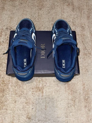 Blå Dior sneakers med vita detaljer - Snygga blå sneakers från Dior med vita och ljusblå detaljer. Skorna har en sportig design med meshpaneler och klassiska snörningar. Dior-logga syns både på innersulan och tungan. Perfekta för dig som vill ha en exklusiv och trendig look.