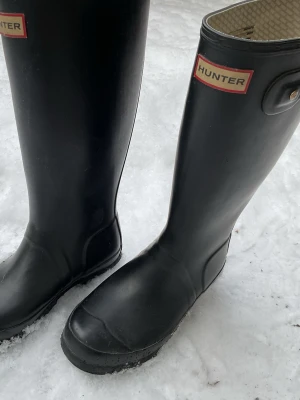 Svarta höga gummistövlar Hunter - Säljer ett par klassiska svarta höga gummistövlar från Hunter. Stövlarna har en justerbar rem upptill och logotypen framtill. Insidan har ett ljust rutigt foder. Perfekta för regniga dagar och snöslask.