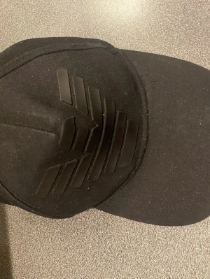 Svart keps från Emporio Armani - Snygg svart keps från Emporio Armani med det klassiska örnlogotypen i svart 3D-tryck framtill. Kepsen har böjd skärm och justerbar baksida. Tillverkad i bomullsmaterial som ger en clean och stilren look.