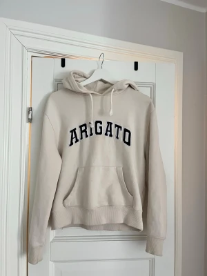 Beige hoodie från Arigato - Snygg beige hoodie från Arigato med svart text framtill. Klassisk modell med huva, dragsnören och stor magficka. Perfekt för en avslappnad och trendig stil. Mjuk bomullskänsla och ribbade muddar vid ärmslut och nederkant.