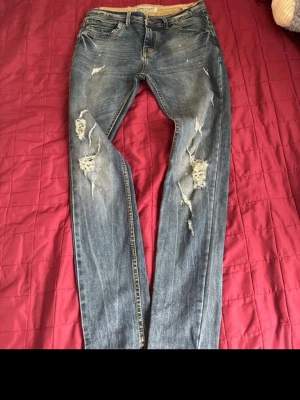 Blå slitna jeans med hål - Säljer ett par blå jeans med slitningar och hål på benen. Jeansen har klassisk femficksmodell, normal midja och raka ben. Materialet är denim i bomull och de har en cool, avslappnad look med tydliga slitningar framtill.