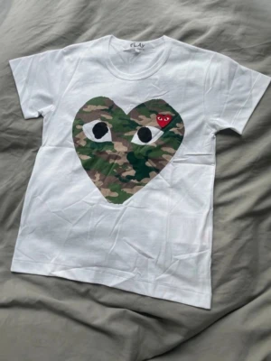 Vit Comme des Garçons Play t-shirt - Vit t-shirt från Comme des Garçons Play med stort hjärta i camouflage-mönster och ögon på bröstet. Liten röd hjärtdetalj med ögon ovanpå. Klassisk passform och rund hals. Perfekt för dig som gillar streetwear och statement-plagg.