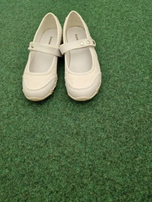 Vita ballerinas med rem från ECCO - Snygga vita ballerinas från graceland med klassisk Mary Jane-rem och två silverfärgade knappar. Skorna har rundad tå, diskreta håldetaljer på sidorna och är tillverkade i mjukt läder. Perfekta för en stilren och bekväm look.