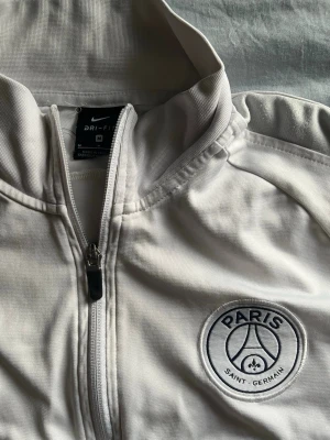 Vit PSG träningsjacka  - Snygg vit träningsjacka från Nike med Paris Saint-Germain-logga på bröstet och röd Nike Swoosh. Jackan har hel dragkedja, ståkrage och två sidofickor. Tillverkad i Dri-FIT-material som andas och passar perfekt till träning eller sport. Köpt på unisport. 