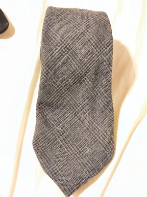 Grå slips från A. Christensen - Snygg grå slips från A. Christensen med diskret rutmönster. Tillverkad i ett mjukt tyg som ger en klassisk och stilren look. Perfekt accessoar för att lyfta din outfit med en elegant touch.