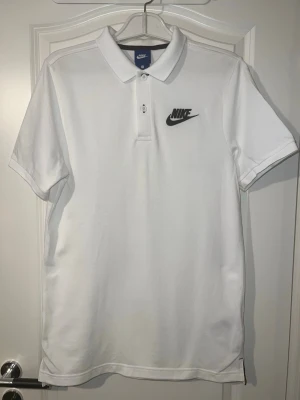 Vit pikétröja från Nike - Klassisk vit pikétröja från Nike med svart broderad logga på bröstet. Tröjan har korta ärmar, krage och knäppning med två knappar framtill. Perfekt för en clean och sportig look. Finns att hämta i Södertälje.