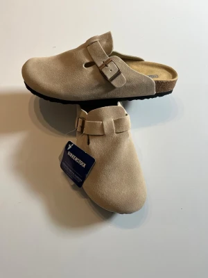 Beige Birkenstock Boston tofflor mocka - Säljer ett par snygga Birkenstock Boston tofflor i beige mocka. De har en klassisk slip-in design med justerbart spänne och bekväm korksula. Perfekta för dig som gillar stilrena och tidlösa skor. Märket syns på innersulan och etiketten sitter kvar.