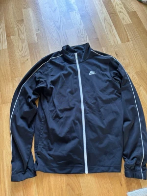 Svart kofta från Nike - Snygg svart kofta från Nike med vit dragkedja och vita detaljer längs ärmarna. Koftan har hög krage, lång ärm och Nike-logga på bröstet. Storlek XS, men passar även bra till storlek S
