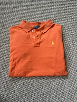 Orange pikétröja från Ralph Lauren - Snygg orange pikétröja från Ralph Lauren med klassisk krage och två knappar framtill. Tröjan har det ikoniska gula broderade logotypen på bröstet och är tillverkad i bomull. Perfekt till golfbanan eller ba ha en stil ren look till sommaren